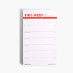 Large designer notepad with light pink background and red lettering, showing a weekly layout on smooth premium paper.